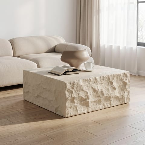 A beige, textured coffee table with a book, cup, and vase on top, in front of a cream-colored couch.